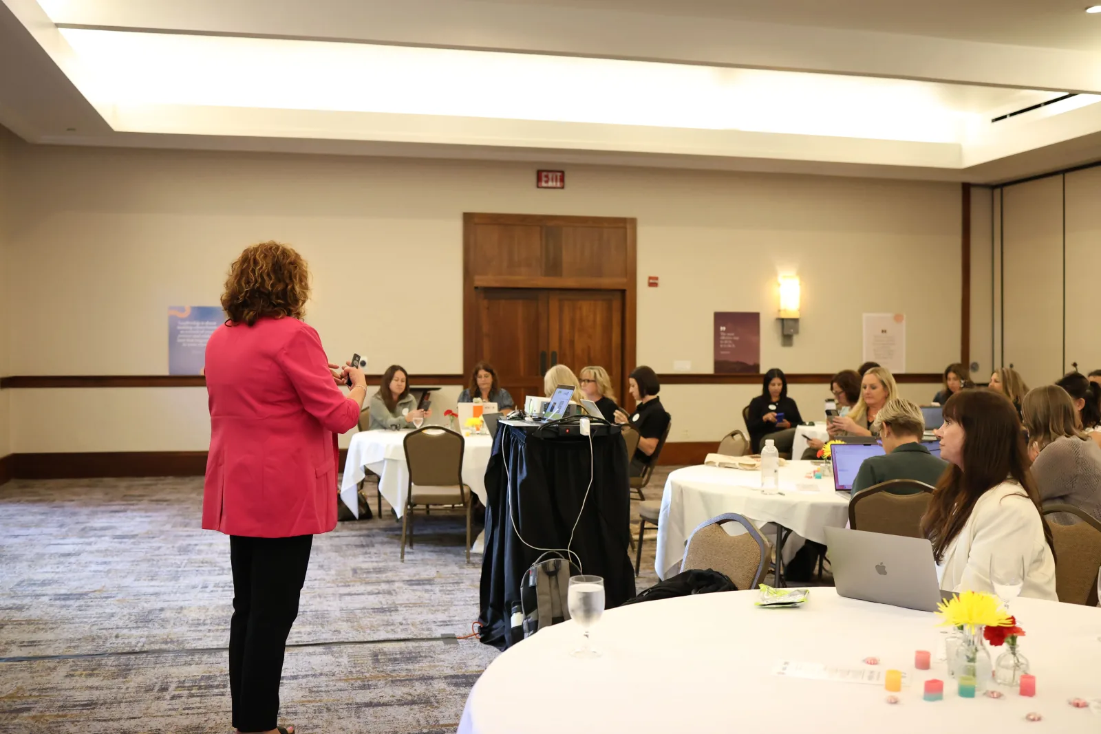 Facilitator addresses a leadership cohort seated with laptops at white-clothed tables